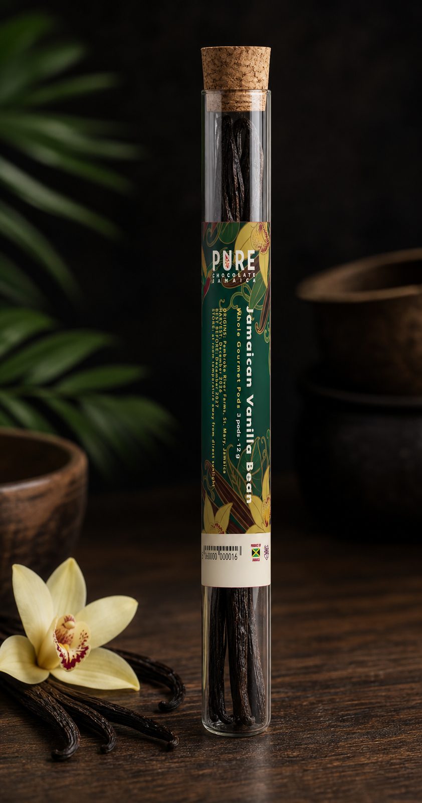 Jamaican Vanilla Bean pods in a corked glass tube with a green orchid-painted wrap, standing on a walnut table beside a vanilla bloom.